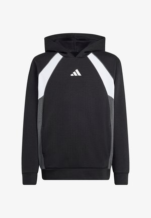 Junge trägt schwarzen und weißen Adidas-Hoodie, Hände in den Taschen, lächelt vor einem einfarbigen hellen Hintergrund.