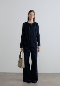 Μαύρη μπλούζα με μακριά μανίκια και πλισέ λαιμόκοψη, συνδυασμένη με σκούρο τζιν φαρδιάς γραμμής. Το μοντέλο κρατάει μια ανοιχτό μπεζ τσάντα χειρός με σκληρή δομή.