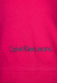 Ljust rosa tyg med en texturerad yta, med inskriptionen "CalvinKleinJeans" i mörkblått längs fållen.