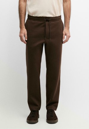 Uomo che indossa pantaloni della tuta marrone scuro con coulisse in vita e sneakers marrone scuro, in piedi davanti a uno sfondo chiaro e uniforme.