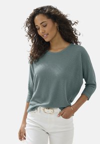 Tropfengrüner, strukturiertes Langarmshirt mit lockerer Passform, kombiniert mit weißen Hosen und einem mit Gold akzentuierten Gürtel. Das Shirt hat einen runden Halsauschnitt.