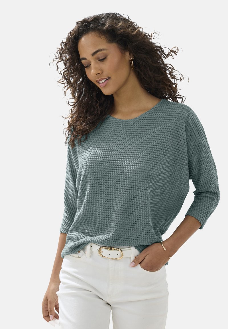 Tropfengrüner, strukturiertes Langarmshirt mit lockerer Passform, kombiniert mit weißen Hosen und einem mit Gold akzentuierten Gürtel. Das Shirt hat einen runden Halsauschnitt.