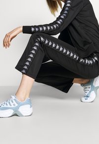 Svart idrottsoverall med vita och ljusblå logodetaljer. Har en hög krage, fickor med dragkedja och korta byxor. Ljusblå sneakers.