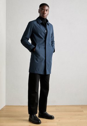 Cappotto impermeabile blu navy con chiusura a singolo bottone, tasche laterali e design elegante. Abbinato a pantaloni neri e stivali alla caviglia.