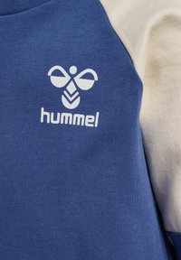 Blauer und cremefarbener Sweatshirt mit einem auffälligen weißen Logo, das ein Bienenmotiv zeigt, und dem Text "hummel" darunter. Weicher Stoff mit einer strukturierten Oberfläche.