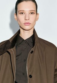 Giacca marrone con colletto testurizzato e camicia nera sotto, con bottoni. Tonalità della pelle liscia e trucco naturale. Sfondo neutro.