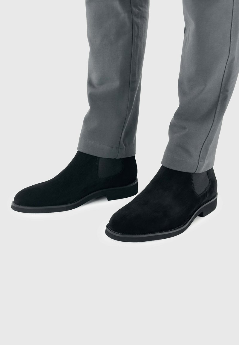 Zwarte suède chelsea boots met elastische zijpanelen en een lage hak, gecombineerd met grijze broeken, tegen een lichtgrijze achtergrond.