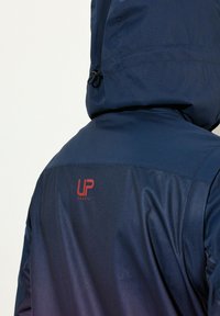 Veste coupe-vent marine avec capuche ajustée, fabriquée en tissu lisse. Présente un logo rouge "UP SPORTS" au dos.