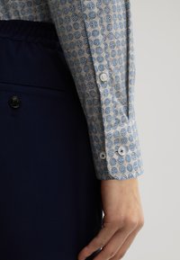 Cuff di una camicia a maniche lunghe a righe blu e bianche accanto a un tessuto blu scuro, con una texture liscia e motivi arrotondati in un design simmetrico.