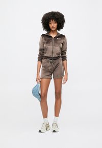 Γυναίκα με σγουρά μαλλιά που φοράει καφέ βελούδινο φούτερ με κουκούλα και σορτς, λευκά αθλητικά παπούτσια, κρατώντας μπλε καπέλο μπέιζμπολ σε απλό φόντο.