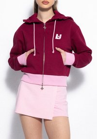 Bordowa bluza z kapturem, zapinana na zamek, z falbaniastym kapturem, różowymi akcentami i logo. W połączeniu z jasnoróżową mini spódnicą o skośnym cięciu.