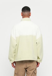 Green and white fleece pullover with a high collar, loose fit, and textured fabric. The design features color blocking and a straight hem.