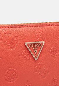 Červená texturovaná kožená peněženka se zvláštními reliéfními vzory a stříbrnou trojúhelníkovou logoplátkou s nápisem "GUESS USA". Zlatý zip na horní straně.