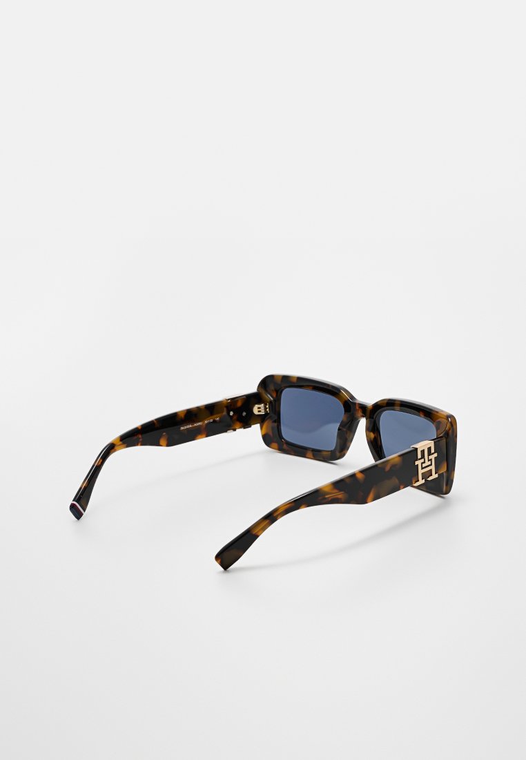 Lunettes de soleil en écaille de tortue avec une monture carrée audacieuse, des verres foncés et un accent en or sur la branche. Texture lisse et design contemporain.