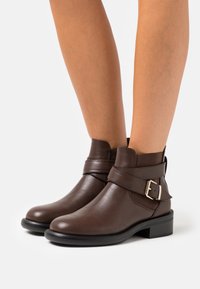 Bottes cheville en cuir marron avec une boucle distincte et un petit talon bloc, offrant un design élégant avec une texture lisse.
