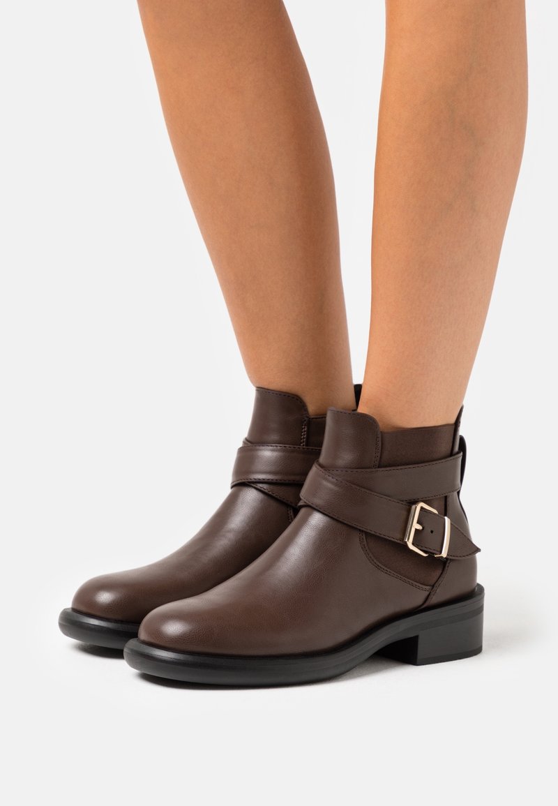 Bottes cheville en cuir marron avec une boucle distincte et un petit talon bloc, offrant un design élégant avec une texture lisse.