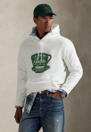Homme portant un sweat à capuche blanc avec un logo de tasse à café verte, chemise à carreaux en dessous, jean, casquette verte, lunettes rondes et montre-bracelet, posant avec la main dans la poche.