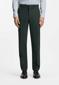 Donkergroene, op maat gemaakte broek met een gestructureerde stof, voorplooien en een rechte snit. Draagt een lichtblauw overhemd en zwarte schoenen.