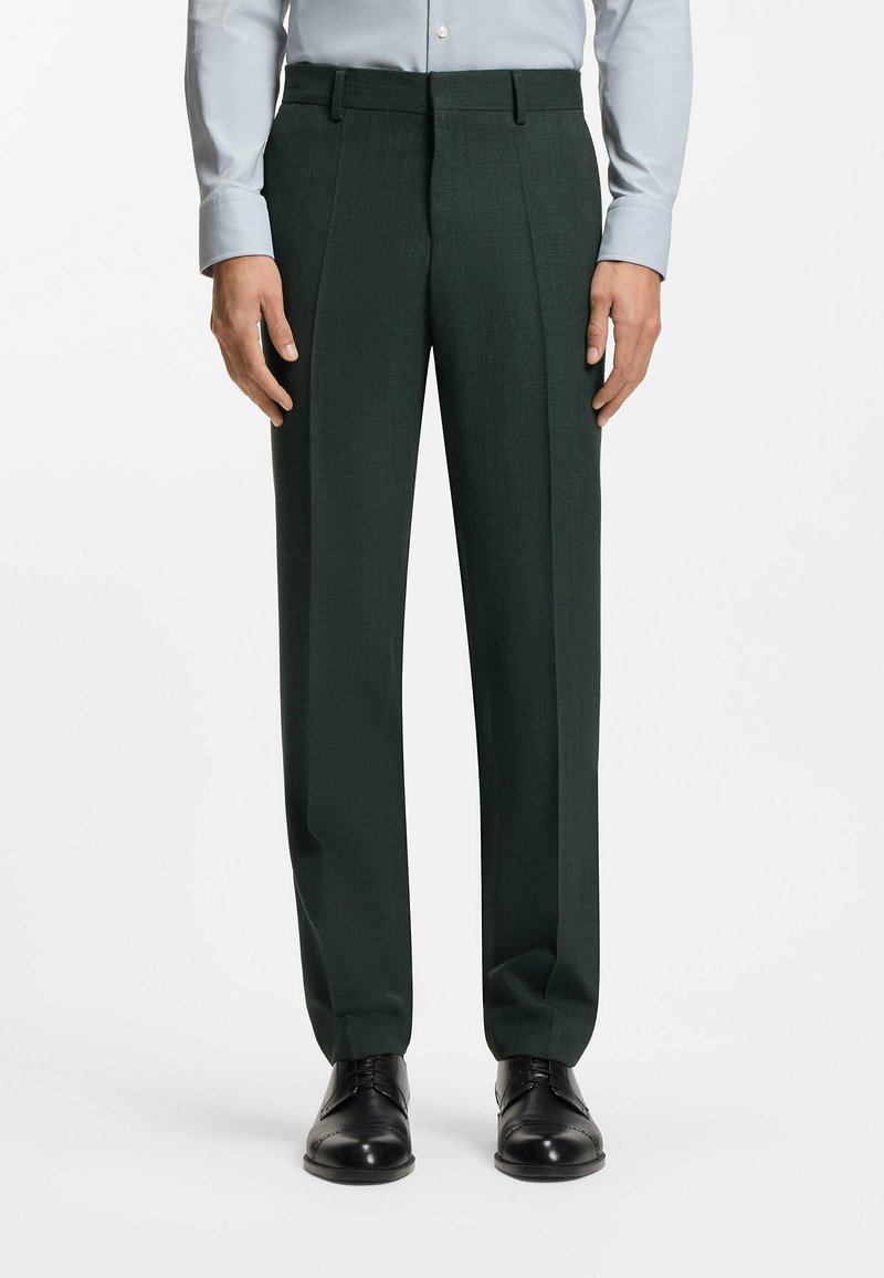 Donkergroene, op maat gemaakte broek met een gestructureerde stof, voorplooien en een rechte snit. Draagt een lichtblauw overhemd en zwarte schoenen.