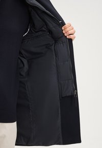 Marineblaues Jacket mit einem glatten, gesteppten Innenfutter. Verfügt über eine Reißverschlusstasche und sichtbare Nähte entlang der Kanten. Hergestellt aus synthetischen Materialien.