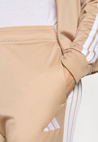 Roka v bežnih trenirkah z belimi črtami in Adidasovim logotipom, v kombinaciji z usklajeno jakno z zadrgo in belo srajco.