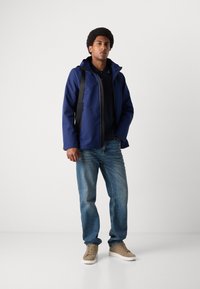 Veste imperméable bleu marine avec capuche, fermeture éclair grise, portée sur un polo noir et un jean bleu clair. Avec des chaussures à lacets beiges.