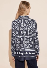 Blusa blu navy con motivi floreali e geometrici bianchi, maniche lunghe, design con colletto e taglio dritto. Tessuto morbido, vestibilità rilassata.