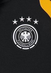 Camisa desportiva preta com um emblema circular. Apresenta um design de águia branca, texto circular e cinco estrelas brancas acima do distintivo.
