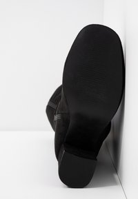 Bottine en daim noir avec une semelle texturée. Présente une finition lisse, une forme légèrement courbée et une fermeture à glissière latérale.