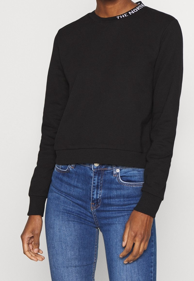Personne portant un sweat-shirt noir à manches longues avec un texte blanc sur le col et un jean en denim bleu sur fond blanc uni.