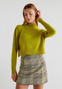 Jasnozielony krótki sweter o prążkowanej fakturze i wysokim dekolcie, w połączeniu z kraciastą spódnicą w odcieniach żółtego, czarnego i białego.