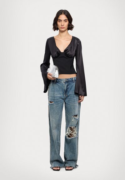 Blusa in satin nero con scollatura a V profonda e maniche a campana, abbinata a jeans di denim blu strappati con grandi buchi. Tiene una clutch argentata.