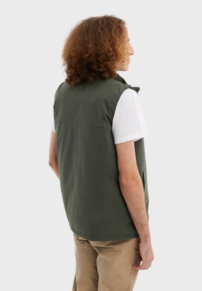 Olivově zelená bezrukávová vesta s hladkou texturou, předními kapsami a vysokým límcem, kombinována s bílým tričkem a béžovými kalhotami.