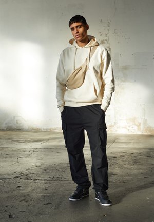 Creme hoodie, sorte cargo bukser og sorte sneakers. Bemærkelsesværdig beige talje taske båret tværs over kroppen. Teksturerede overflader, afslappet pasform, minimalistisk design.