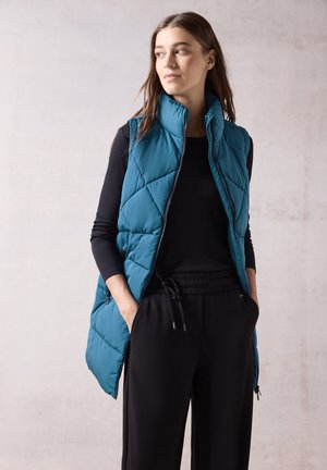 Gilet isolant teal avec un motif en losanges, col haut et poches latérales, porté sur une chemise noire à manches longues et un pantalon noir.