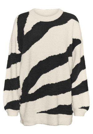 Person iført en fluffet hvid sweater med sorte abstrakte striber, sorte kunstlæderbukser med forreste slidser og sort-hvide sneakers.