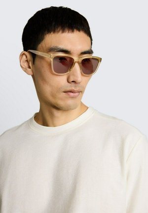 Homme portant des lunettes de soleil beiges translucides et un t-shirt blanc à col rond, légèrement tourné vers la gauche sur un fond clair uni.