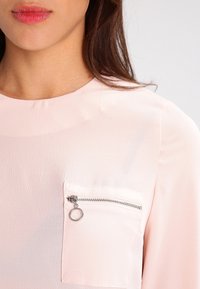 Light pink blouse featuring a textured fabric, round neckline, and a chest pocket with a silver zip and circular hardware detail.
