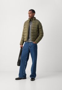 Oliven grøn pufferjakke, quiltet design, båret over en grå strikketrøje. Løse blå jeans og sorte sko, holder en sort skuldertaske.