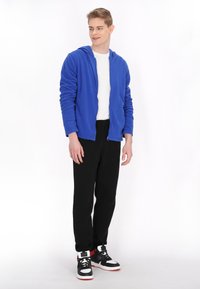 Felpa blu con zip, t-shirt bianca sotto, pantaloni neri e sneakers nere con dettagli bianchi e rossi. Tessuto morbido, vestibilità casual.