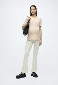 Haut en maille rose clair, associé à un pantalon évasé crème. Le mannequin tient un sac en cuir noir et porte des chaussures noires épaisses. Design simple et épuré.