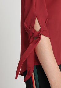 Blusa bordeaux a maniche lunghe con legature annodate ai polsini, caratterizzata da una texture leggera e liscia e un sottile dettaglio con spacco.