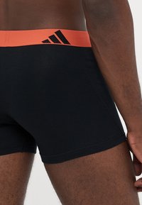 Črne bokserice z oranžnim elastičnim pasom, ki vsebuje črni adidas logotip, z bližnjim pogledom na zadnjo levo stran.