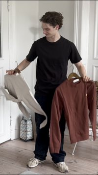 Fekete póló, világos bézs hosszúujjú ing és rozsdabarna pulóver, amelyet egy személy tart, a háttérben fás akasztók és egy mintás lámpás látható.