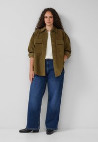 Oliwkowa kurtka koszulowa z korduroyu z dwoma kieszeniami na piersi, zestawiona z szerokimi niebieskimi dżinsami i czarnymi botkami za kostkę.