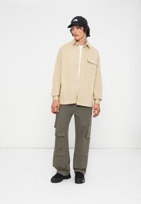 Giovane uomo che indossa una camicia beige a bottoni, pantaloni cargo verdi oliva, sneakers nere e un cappellino nero, in piedi su uno sfondo bianco uniforme.
