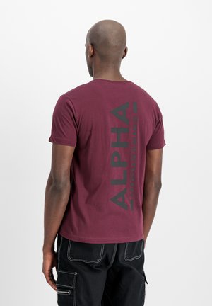 Camiseta burdeos de algodón con cuello redondo, que presenta el texto vertical en negro "ALPHA INDUSTRIES" en la parte de atrás. Mangas cortas, corte estándar.