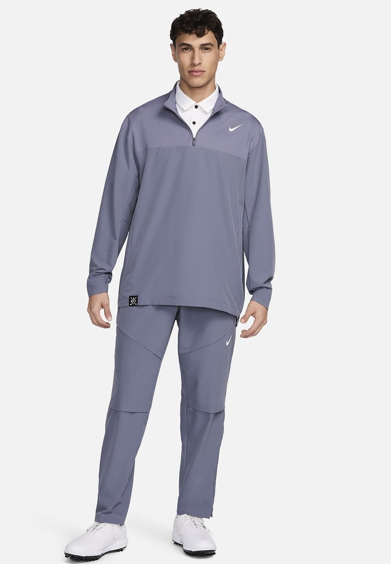Suéter gris de mangas largas con media cremallera y cuello blanco, combinado con pantalones gris a juego que presentan paneles geométricos y un pequeño logo.