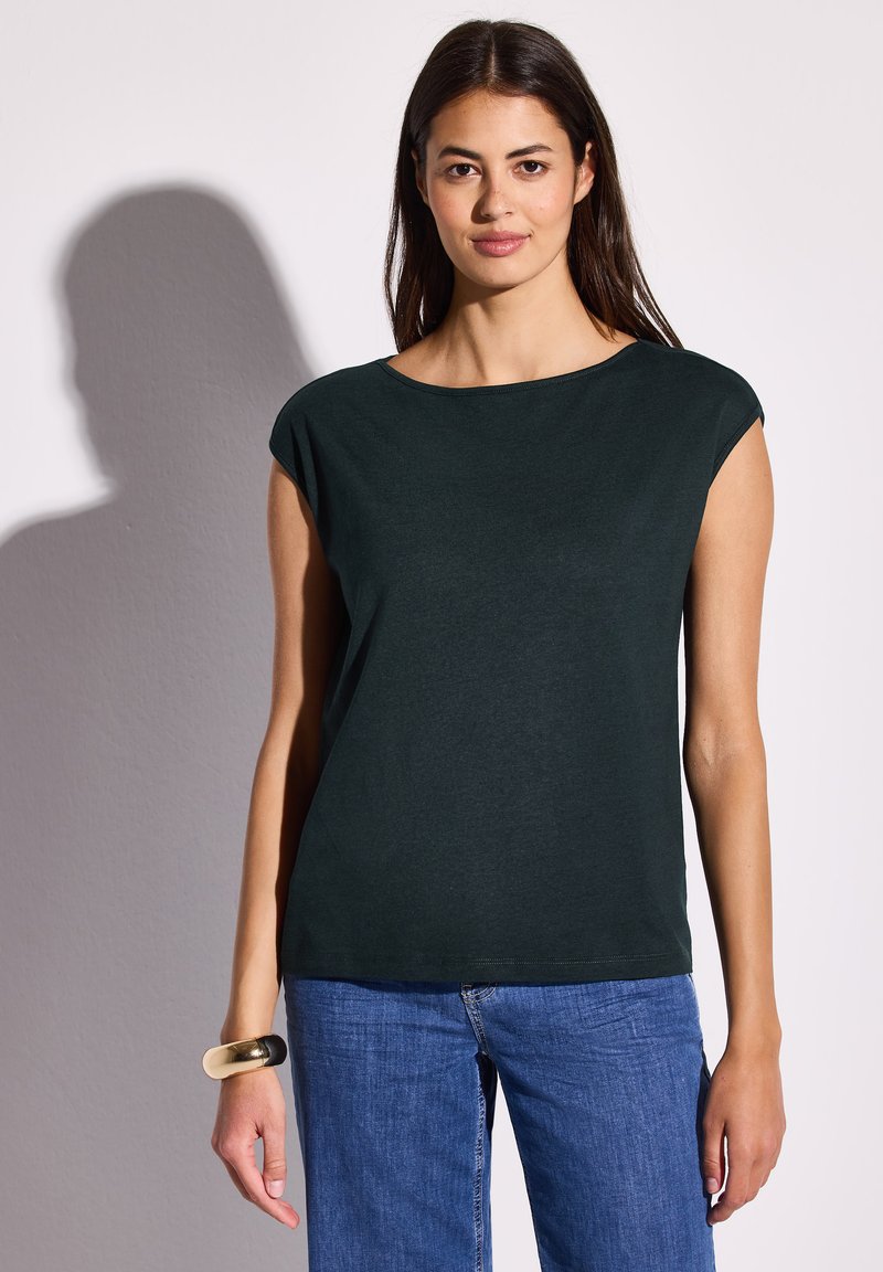 Tummanvihreä, lyhythihainen paita, jossa on leveä kaula-aukko ja rento istuvuus. Valmistettu pehmeästä materiaalista, yhdistetty vaaleansinisiin farkkuihin. Yksinkertainen muotoilu.