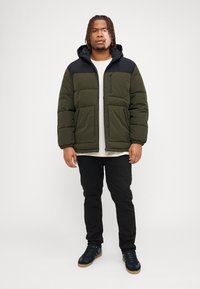Doudoune vert olive avec capuche et épaules noires, deux poches avant et une poche poitrine zippée, portée sur une chemise légère et un pantalon noir.